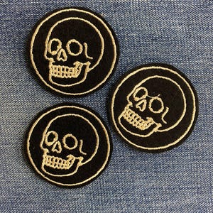 May include: Three black circular patches with a skull design. Each patch features a white embroidered skull within a white circular border. The patches are arranged on a blue denim background.