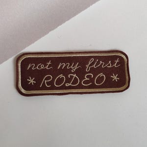 May include: Brown felt patch with a gold embroidered border. The text "not my first *RODEO*" is embroidered in gold.