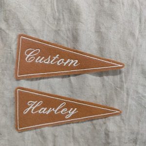 May include: Two brown felt pennants with white embroidered text. One pennant reads "Custom" and the other reads "Harley". Each pennant has a white border. The pennants are triangular in shape and are displayed on a neutral-coloured fabric background.