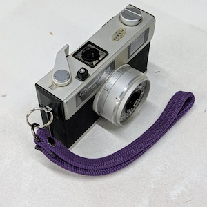May include: A vintage Canonet 28 camera with a silver lens and black body. The camera has a purple wrist strap attached. The lens has a silver ring with numbers and markings. The camera is labelled "PASSED".