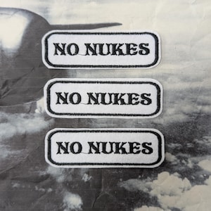 Może przedstawiać: Trzy prostokątne naszywki z napisem "NO NUKES" wyhaftowanym na czarno na białym tle. Każda naszywka ma czarną obwódkę. Tło to czarno-biały obraz chmur i samolotu.
