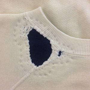 May include: A white sweatshirt with a navy blue patch sewn into the neckline. The patch has a jagged edge and is slightly frayed.