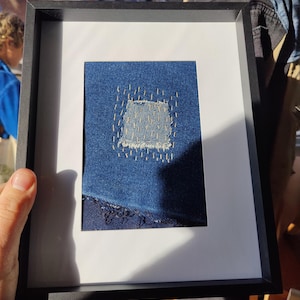 Puede incluir: Una obra de arte enmarcada que presenta un parche de mezclilla azul con un agujero que se ha reparado con costuras. La mezclilla est&aacute; deshilachada en los bordes.