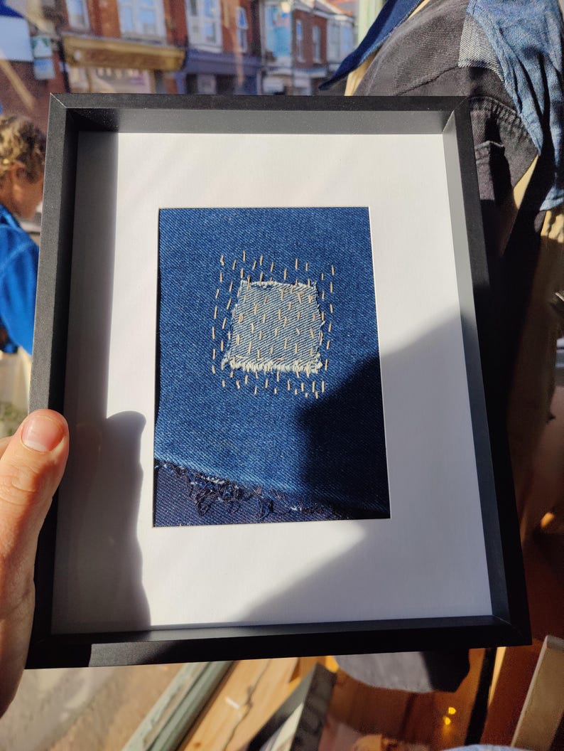Puede incluir: Una obra de arte enmarcada que presenta un parche de mezclilla azul con un agujero que se ha reparado con costuras. La mezclilla est&aacute; deshilachada en los bordes.