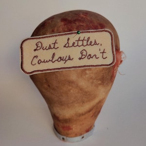 May include: A tan fabric mannequin head with a brown and white embroidered patch that reads "Dust settles. Cowboys Don't."