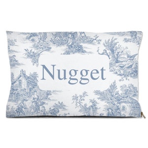 May include: A white and blue toile patterned dog bed with a black border and a zipper. The bed has a white rectangle with the name "Nugget" in blue text.