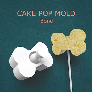 May include: A white bone-shaped cake pop mold next to a cake pop on a stick. The cake pop is yellow and bone-shaped. The text "CAKE POP MOLD Bone" is in the top center of the image.
