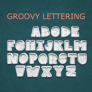 May include: White plastic alphabet cookie cutters with decorative designs. The letters are arranged in rows against a teal background. The words "GROOVY LETTERING" are written in orange above the letters.