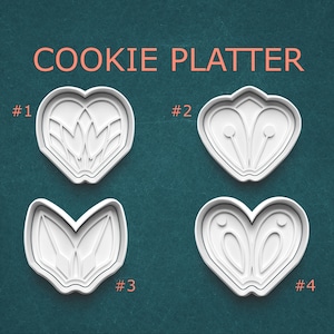 May include: Four white plastic cookie cutters in heart shapes with different floral and abstract designs. The text "COOKIE PLATTER" is in orange above the cutters, with numbers #1 through #4 next to each cutter.