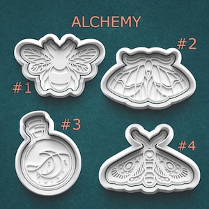 May include: Four white plastic cookie cutters with an alchemy theme. The cutters are shaped like a bee, a moth, a potion bottle with a moon and stars, and a moth with star and circle details. The word "ALCHEMY" is printed at the top.