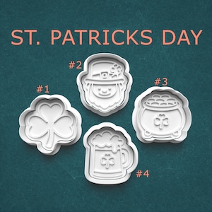 May include: Four white plastic cookie cutters for St. Patrick's Day. The shapes include a shamrock, a leprechaun face, a pot of gold, and a beer mug with a shamrock. The words "ST. PATRICKS DAY" are in orange text at the top.