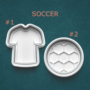 May include: Two white plastic cookie cutters. One is shaped like a soccer jersey, and the other is a soccer ball. The word "SOCCER" is printed in orange above the cookie cutters. The number "#1" is printed next to the jersey cutter, and "#2" is next to the ball cutter.