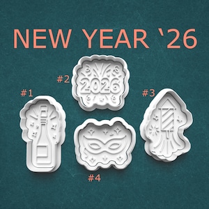 May include: Four white plastic cookie cutters with a New Year's theme. The cutters include a champagne bottle, the number 2026, a masquerade mask, and a firework. The words "NEW YEAR '26" are in orange text above the cutters.