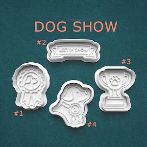 May include: Four white plastic cookie cutters in the shape of dog show-themed designs. The designs include a ribbon, a banner that says "BEST IN SHOW", a trophy with a paw print, and a dog with a ribbon.