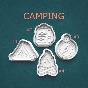May include: Four white camping-themed cookie cutters: a tent, backpack, compass, and campfire. The word "CAMPING" is written in coral-colored letters above the cutters. Each cutter is labeled with a number.