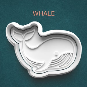 May include: A white whale-shaped cookie cutter with the word "WHALE" in orange above it. The cutter has a detailed design, including the whale's tail, body, and belly lines. The cutter is on a teal background.
