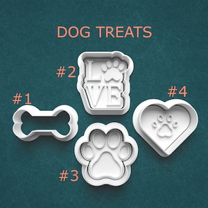 May include: Four white plastic cookie cutters for dog treats. The shapes include a bone, the word "LOVE" with a paw print, a paw print, and a heart with a paw print. The words "DOG TREATS" are in orange text at the top.