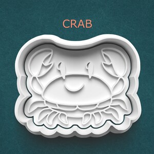 May include: White plastic crab-shaped cookie cutter with a smiling face and claw details. The word "CRAB" is printed in orange above the cutter. The cutter has a scalloped edge and is set against a teal background.
