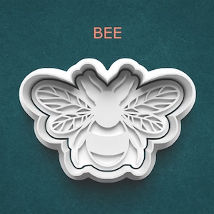 May include: White bee-shaped cookie cutter with detailed wing and body patterns. The word "BEE" is printed in orange above the cutter. The cutter is on a teal background.