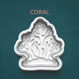 May include: White coral-shaped cookie cutter with the word "CORAL" in orange. The cutter has a layered design, with the coral's intricate branches and base clearly defined. The background is a teal color.