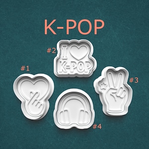 May include: Four white plastic cookie cutters with K-POP themes. Designs include a heart with fingers, a heart with "I K-POP" text, a peace sign with a star, and headphones. The word "K-POP" is in orange.