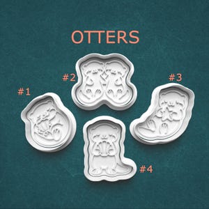 May include: Four white plastic cookie cutters labeled OTTERS with numbers #1 through #4. Each cutter features a different otter design: one holding a shell, two holding hands, one holding a star, and one standing while holding a shell.