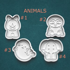 May include: Four white plastic animal-shaped cookie cutters: a rhino, a sloth, a seal, and an elephant. The word "ANIMALS" is printed in orange above the cutters, with numbers identifying each animal. The cutters are on a teal background.