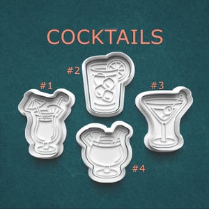 May include: Four white plastic cocktail-shaped cookie cutters. The word "COCKTAILS" is printed in orange above the cutters. The cutters are numbered 1 through 4. The cutters are shaped like a cocktail with a straw and umbrella, a glass with ice and a lemon slice, a martini, and a cocktail.