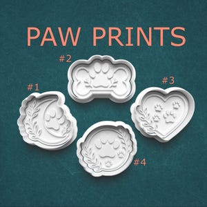 May include: Four white plastic cookie cutters with paw print designs. The cutters are in the shapes of a crescent moon, a bone, a heart, and a circle. The words "PAW PRINTS" are in orange letters at the top.