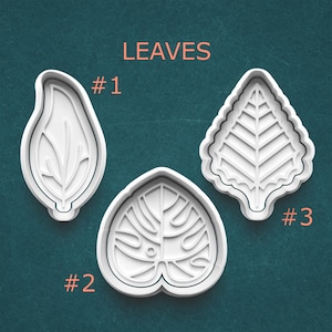 May include: Three white plastic leaf-shaped cookie cutters. The word "LEAVES" is printed in orange above the cutters. Each cutter is labeled with a number: #1, #2, and #3. The background is a teal color.