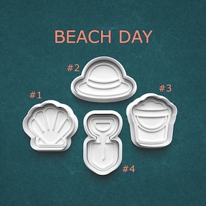 May include: Four white plastic cookie cutters in beach-themed shapes: a seashell, a sun hat, a bucket, and a shovel. The words "BEACH DAY" are printed in orange at the top of the image.
