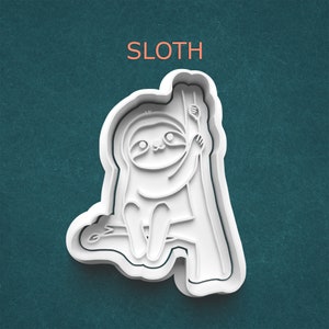 May include: White plastic cookie cutter in the shape of a cartoon sloth hanging from a tree branch. The word "SLOTH" is printed in orange above the sloth. The cutter has a detailed design with a cute, smiling sloth.