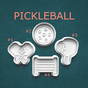 May include: White pickleball-themed cookie cutters. The image shows four different cookie cutter designs: a paddle and ball, a ball, a paddle, and a net. The word "PICKLEBALL" is written in orange letters at the top.