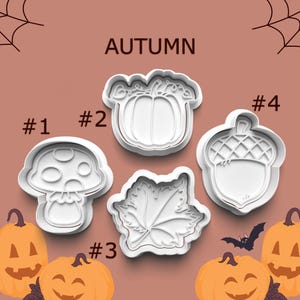 May include: Set of five white plastic cookie cutters shaped like an acorn, a mushroom, a pumpkin, a leaf, and a maple leaf. The cutters are labeled with numbers 1 through 4. The word "AUTUMN" is written above the cutters.