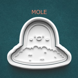 May include: A white plastic cookie cutter in the shape of a cartoon mole emerging from the ground. The word "MOLE" is printed in orange above the cookie cutter on a teal background.