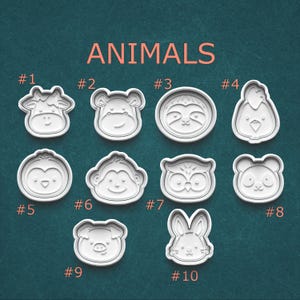 May include: A collection of ten white plastic animal-shaped cookie cutters. The cutters include a cow, bear, sloth, chicken, penguin, monkey, owl, panda, pig, and rabbit. The word "ANIMALS" is printed in orange above the cutters.