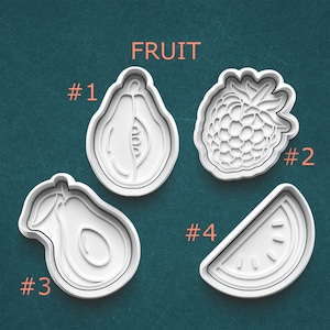 May include: Four white plastic fruit-shaped cookie cutters: a papaya, a raspberry, an avocado, and a watermelon slice. The word "FRUIT" is above the cutters, with numbers next to each fruit.