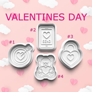 May include: White Valentine's Day cookie cutters in various shapes: a heart bullseye, a phone with a heart and the word "SEND", a heart-shaped lock, and a teddy bear holding a heart. The text "VALENTINES DAY" is displayed in pink.