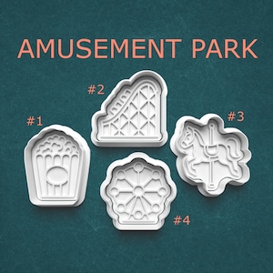 May include: Four white plastic cookie cutters in the shapes of amusement park icons. The shapes include popcorn, a roller coaster, a carousel horse, and a Ferris wheel. The words "AMUSEMENT PARK" are printed in coral.