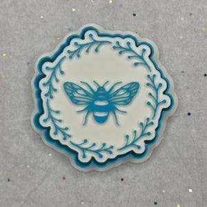 May include: A white and blue decorative bee-themed item. The design features a blue bee in the center, surrounded by a blue floral border. The item has a scalloped edge and is set against a light background.