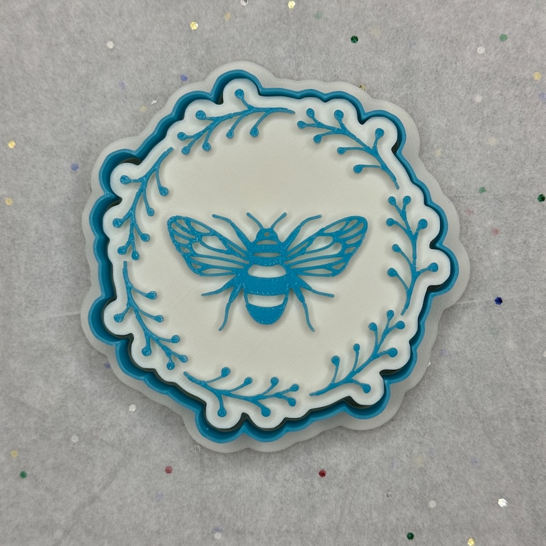 May include: A white and blue decorative bee-themed item. The design features a blue bee in the center, surrounded by a blue floral border. The item has a scalloped edge and is set against a light background.