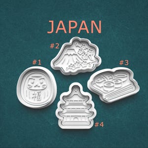 May include: Four white plastic cookie cutters with Japanese themes. The cutters include a Daruma doll, Mount Fuji with cherry blossoms, sushi, and a pagoda. The word "JAPAN" is written in orange at the top.
