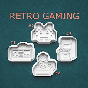 May include: Four white plastic cookie cutters in retro gaming shapes. The shapes include a game controller, a space invader, an arcade machine, and a joystick. The words "RETRO GAMING" are written in orange at the top.