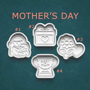 May include: Four white plastic cookie cutters for Mother's Day. The cutters include a mother and child, a gift box with a heart, a flower and heart, and a trophy that says "Best Mom". The words "Mother's Day" are above the cutters.