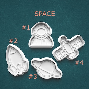 May include: White plastic cookie cutters in space-themed shapes: an astronaut, a rocket, a planet with rings, and a satellite. The word "SPACE" is printed in orange above the cutters. Numbers are printed next to each cutter.