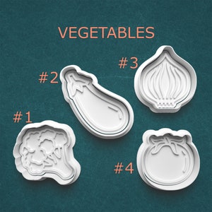 May include: Four white plastic vegetable-shaped cookie cutters. The cutters are shaped like a cauliflower, eggplant, onion, and tomato. The word "VEGETABLES" is printed in orange above the cutters. Numbers 1-4 are printed next to each cutter.