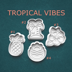 May include: Four white plastic cookie cutters with tropical designs: a pineapple, a palm tree, a hibiscus flower, and a sunset over the ocean. The text "TROPICAL VIBES" is above the cutters.