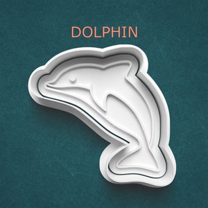 May include: A white dolphin-shaped cookie cutter with the word "DOLPHIN" in orange text above it. The cutter has a three-dimensional design and is set against a teal background.