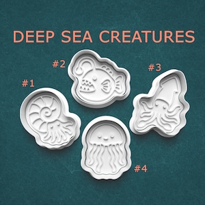 May include: Four white plastic cookie cutters labeled DEEP SEA CREATURES. Number 1 is an ammonite, number 2 is an anglerfish, number 3 is a squid, and number 4 is a jellyfish. Each cutter features a raised outline and internal details for imprinting.