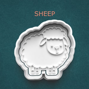 May include: A white plastic sheep-shaped cookie cutter with the word "SHEEP" in orange letters above it. The cutter has a detailed outline of a sheep with a fluffy body and a smiling face. The background is a teal color.